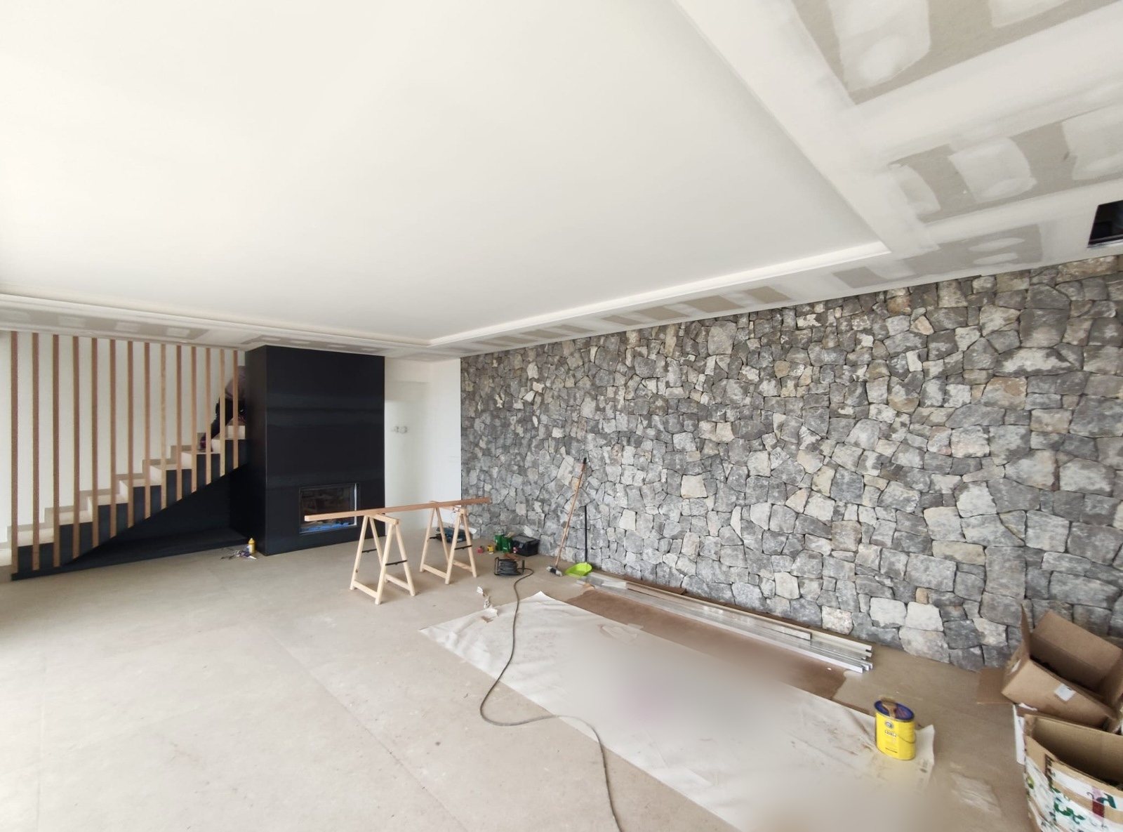 Open-plan living area under construction with a stone feature wall, wood-slat stair screen and black fireplace